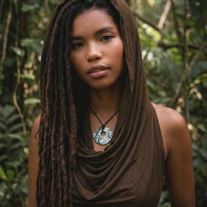 May include: A woman wearing a brown cowl neck top and a matching hood. She has long dreadlocks and a necklace with a circular pendant. The pendant has a blue, green, and white design. The background is a lush green forest.