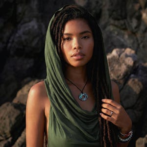 Peut inclure: Une femme porte un haut à col roulé vert foncé et une capuche assortie. Elle a de longues dreadlocks et un collier pendentif avec un motif circulaire bleu et vert. Elle porte des bracelets et une bague.