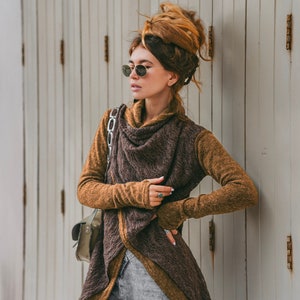 Puede incluir: Una mujer lleva un suéter de punto marrón y tostado con un diseño largo y fluido. Lleva pantalones vaqueros grises y gafas de sol redondas. Está de pie frente a una pared de madera blanca.