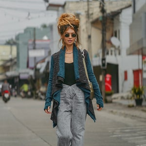 Puede incluir: Una mujer con un cárdigan azul y gris, un top negro, pantalones vaqueros grises y botas negras. Está caminando por la calle.