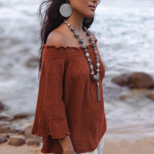 Puede incluir: Una mujer con un top color óxido con hombros descubiertos y mangas acampanadas, combinado con pantalones color crema. Está adornada con pendientes grises redondos grandes, un collar de cuentas y una pulsera plateada. El fondo muestra una playa y el océano.