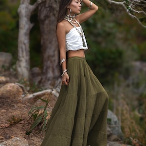 May include: A woman wearing a white tube top and a long olive green skirt. She is adorned with layered shell necklaces and silver bracelets. The outfit is suitable for a bohemian or resort style.