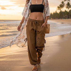 May include: A woman wearing a black crop top, brown pants, and a floral kimono walks on a beach. She is wearing a straw hat, sunglasses, and carrying a woven bag. The pants have a decorative design on the side.