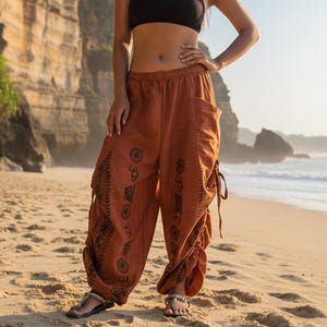 May include: A person wearing a black crop top and rust-coloured harem trousers with a decorative print. The trousers have pockets and drawstring details. The person is standing on a sandy beach with the ocean in the background.