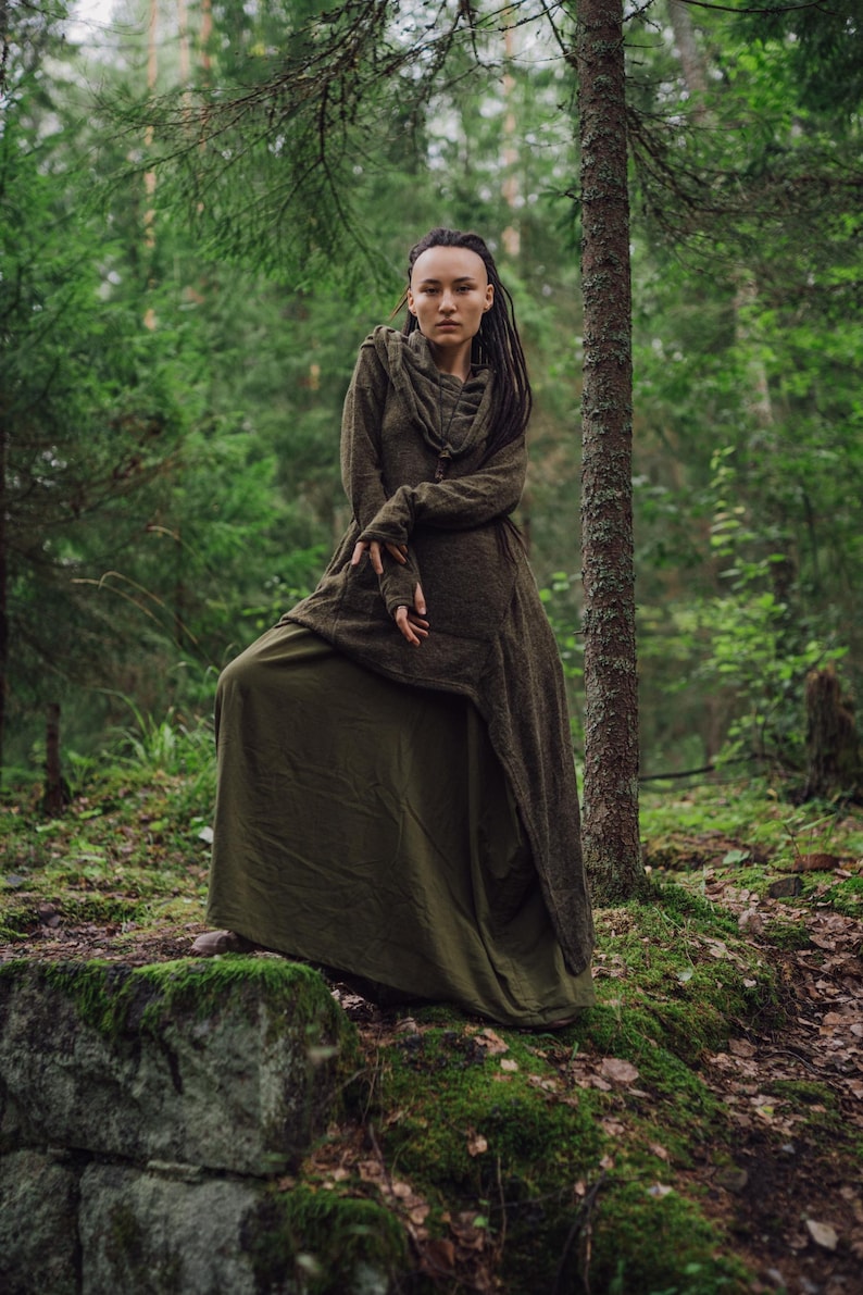 May include: A woman wearing a long, dark green hooded dress stands in a forest. She has dreadlocks and is looking directly at the camera.