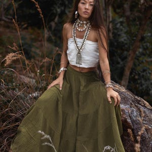 May include: A woman wearing a white tube top and a long olive green tiered skirt. She is adorned with shell necklaces, bracelets, and earrings. The background features a natural outdoor setting with trees and foliage.