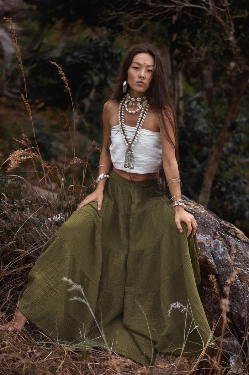 May include: A woman wearing a white tube top and a long olive green tiered skirt. She is adorned with shell necklaces, bracelets, and earrings. The background features a natural outdoor setting with trees and foliage.