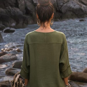 May include: An olive green top with a textured, crinkled fabric. The top has a round neckline and three-quarter sleeves with button details. The person is wearing silver bracelets and earrings. The background is a rocky shoreline with water.