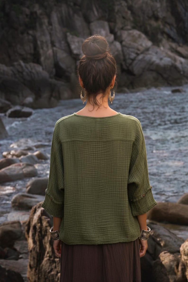 May include: An olive green top with a textured, crinkled fabric. The top has a round neckline and three-quarter sleeves with button details. The person is wearing silver bracelets and earrings. The background is a rocky shoreline with water.