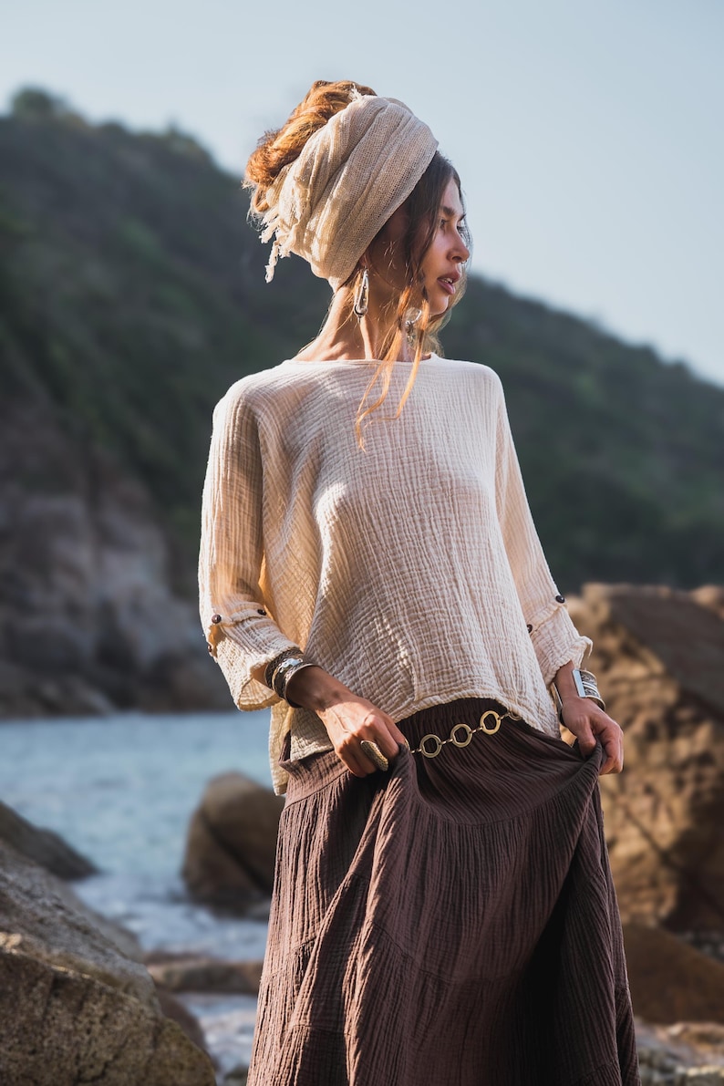 May include: A cream-coloured long-sleeved top with a textured weave, worn with a brown skirt. The outfit features a gold-toned belt and silver bracelets. The model is wearing a headscarf and earrings, with a natural outdoor backdrop.