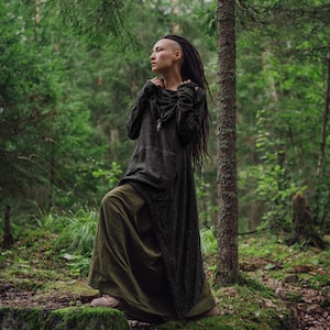 May include: A woman with dreadlocks wears a long, dark green hooded tunic with a long skirt. She stands in a forest with green trees and moss on the ground.