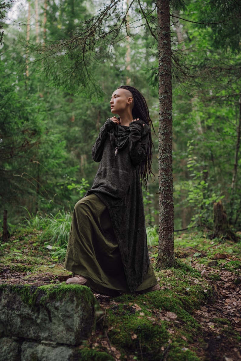 May include: A woman with dreadlocks wears a long, dark green hooded tunic with a long skirt. She stands in a forest with green trees and moss on the ground.