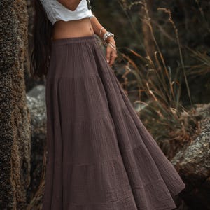 May include: A long, tiered skirt in a muted brown color. The skirt is made of a textured fabric and has a flowing, bohemian style. The model is wearing a white top and jewelry.