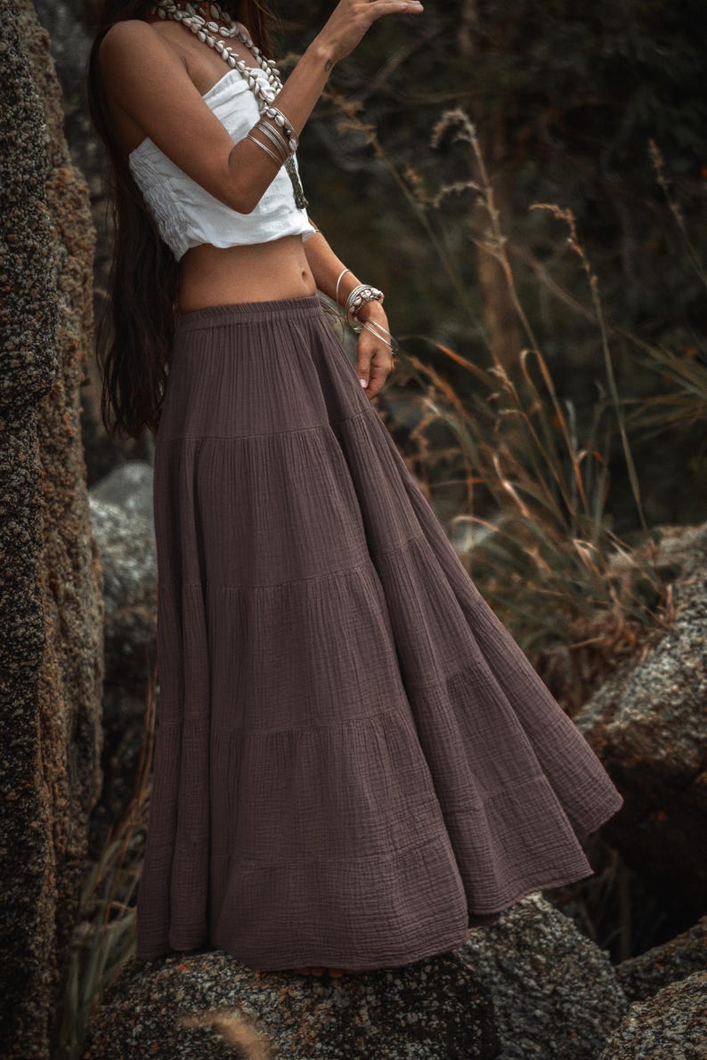 May include: A long, tiered skirt in a muted brown color. The skirt is made of a textured fabric and has a flowing, bohemian style. The model is wearing a white top and jewelry.