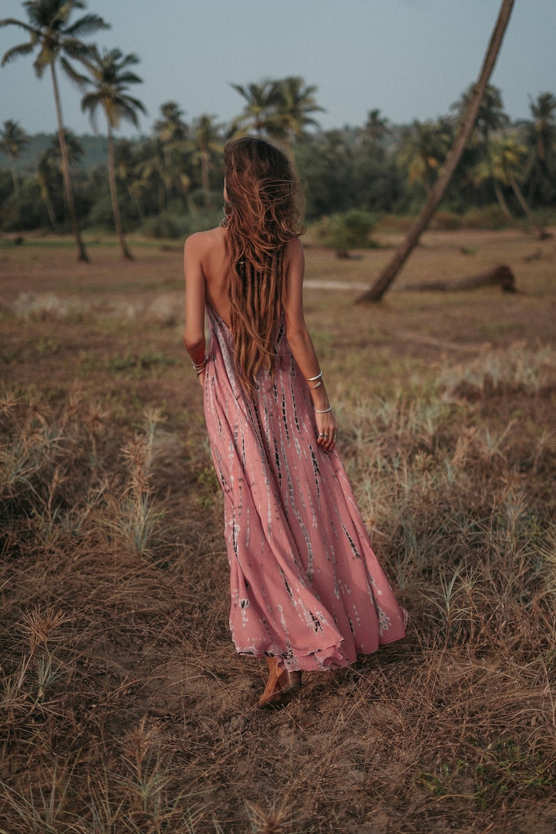 May include: A woman wearing a pink and white tie-dye maxi dress with long brown hair, walking through a field of tall grass.