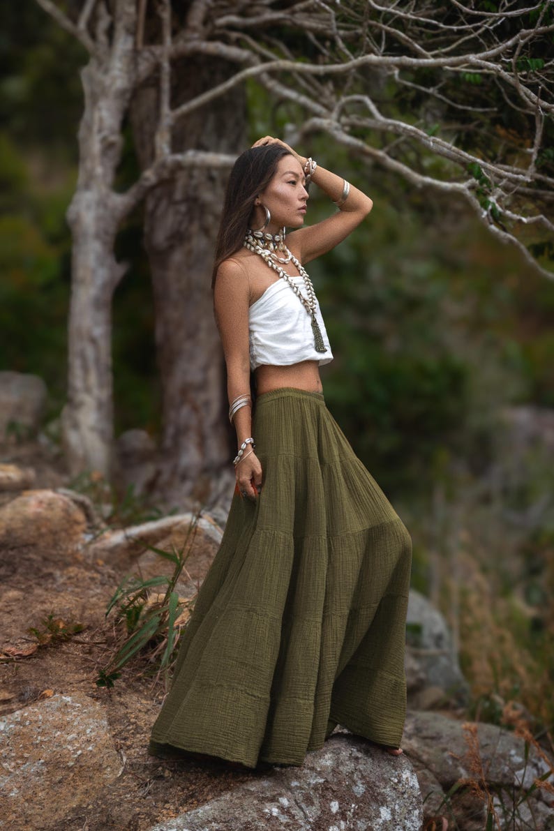 May include: A woman wearing a white tube top and a long olive green skirt. She is adorned with layered shell necklaces and silver bracelets. The outfit is suitable for a bohemian or resort style.