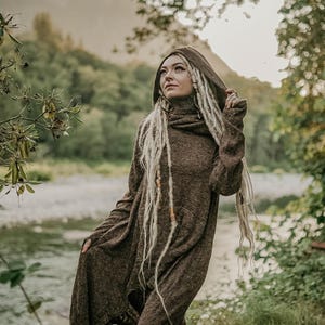 May include: A person wearing a brown hooded sweater with long, blonde dreadlocks. The person is standing in a forest setting with a river in the background.
