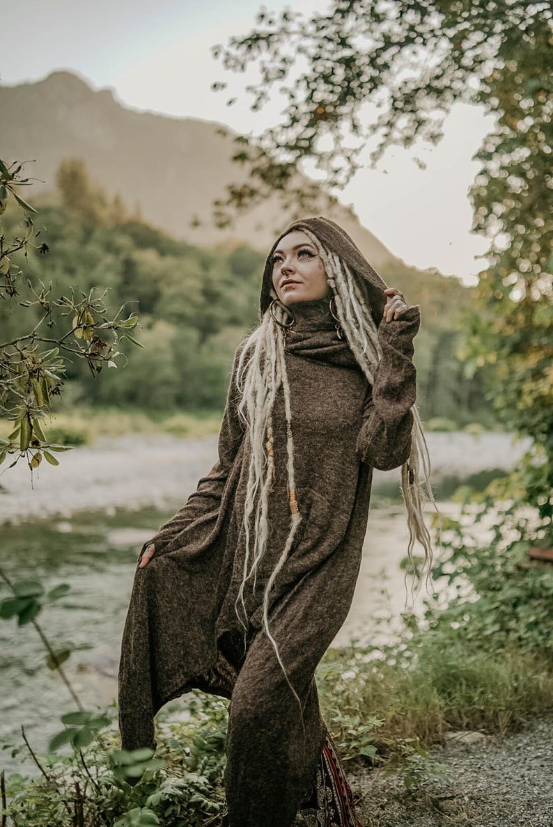 May include: A person wearing a brown hooded sweater with long, blonde dreadlocks. The person is standing in a forest setting with a river in the background.