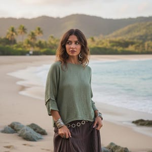 May include: A woman on a beach wearing a sage green top and a brown skirt. The top has button details on the sleeves. The skirt is long and flowing. Silver bracelets and a belt with circular links complete the outfit.