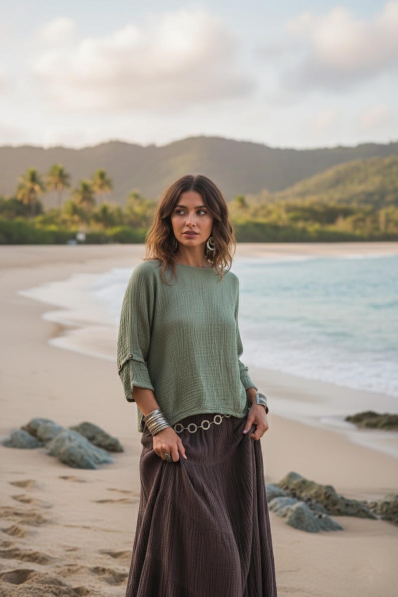 May include: A woman on a beach wearing a sage green top and a brown skirt. The top has button details on the sleeves. The skirt is long and flowing. Silver bracelets and a belt with circular links complete the outfit.