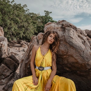 May include: A woman wearing a yellow maxi dress with a white belt and a silver pendant necklace. She is sitting on a rock with a cloudy sky in the background.