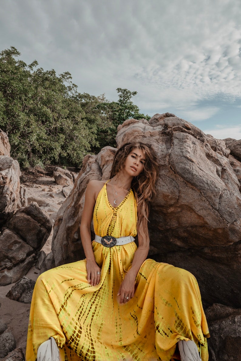 May include: A woman wearing a yellow maxi dress with a white belt and a silver pendant necklace. She is sitting on a rock with a cloudy sky in the background.