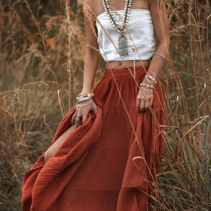 May include: A woman wearing a white tube top and a rust-colored tiered skirt. She is adorned with shell necklaces, silver bracelets, and a pendant necklace. The photo is taken outdoors in a field of tall, dry grass.