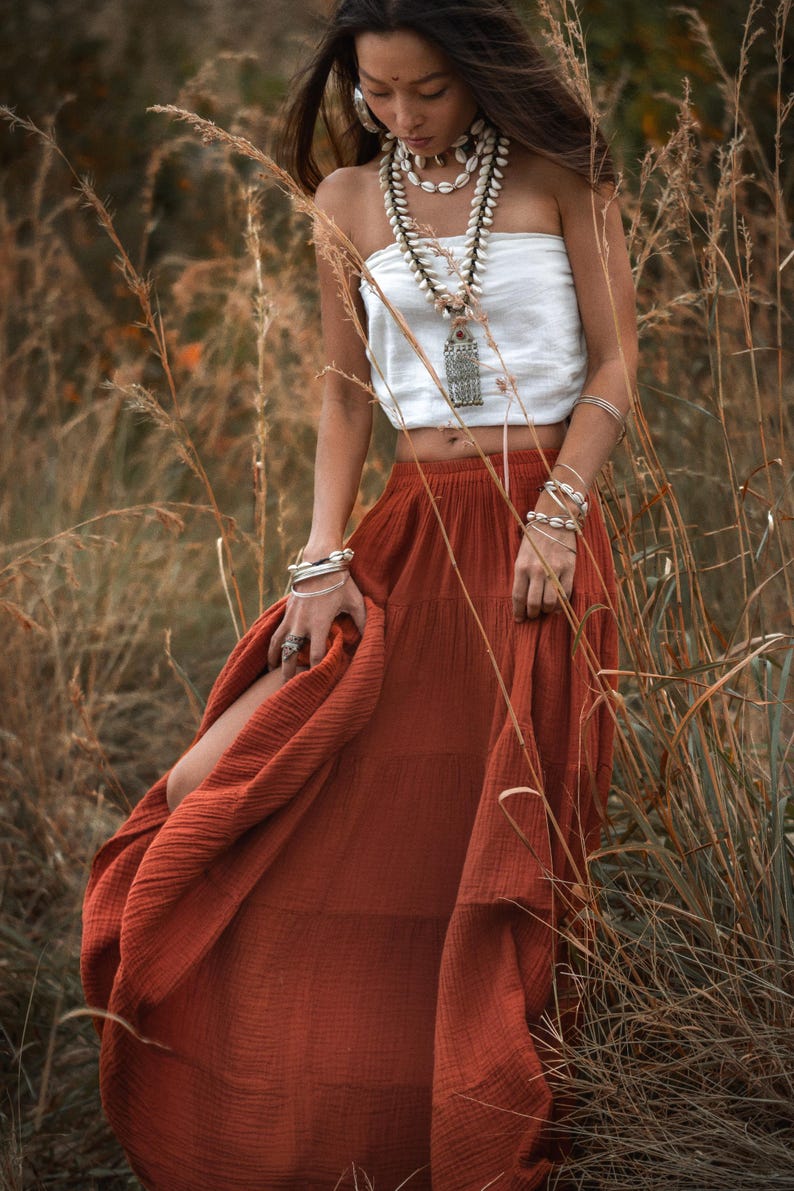 May include: A woman wearing a white tube top and a rust-colored tiered skirt. She is adorned with shell necklaces, silver bracelets, and a pendant necklace. The photo is taken outdoors in a field of tall, dry grass.