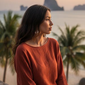 May include: A rust-coloured long-sleeved top with a textured, ribbed design. The top features a round neckline and buttoned cuffs. The image is set against a scenic backdrop of palm trees, the ocean, and mountains.
