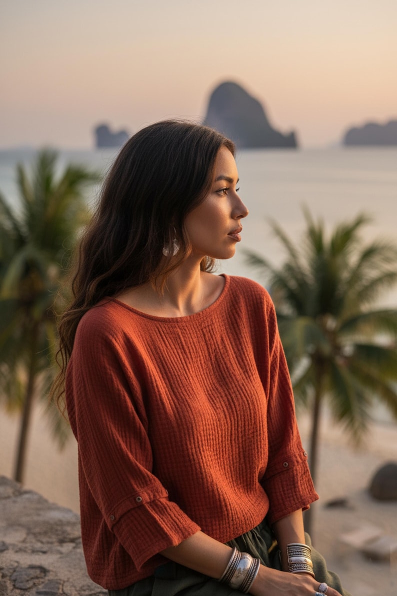 May include: A rust-coloured long-sleeved top with a textured, ribbed design. The top features a round neckline and buttoned cuffs. The image is set against a scenic backdrop of palm trees, the ocean, and mountains.