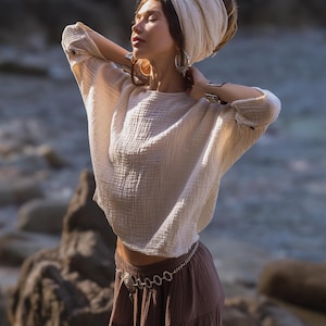 May include: A cream-coloured, long-sleeved top with a textured, crinkled appearance. The top is paired with a brown skirt and a silver chain belt. The model is wearing a cream-coloured head wrap and large silver hoop earrings. The background is a blurred outdoor scene.