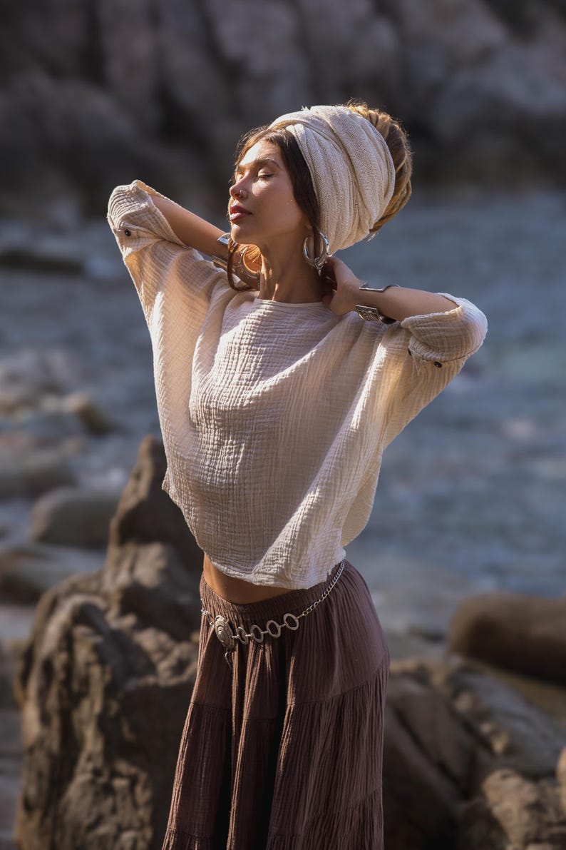 May include: A cream-coloured, long-sleeved top with a textured, crinkled appearance. The top is paired with a brown skirt and a silver chain belt. The model is wearing a cream-coloured head wrap and large silver hoop earrings. The background is a blurred outdoor scene.