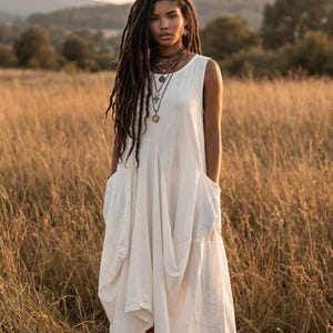 Puede incluir: Un vestido sin mangas de color blanco roto con bolsillos grandes. El vestido lo lleva una persona con rastas largas y oscuras, de pie en un campo de hierba alta y seca. La persona lleva varios collares y sandalias.
