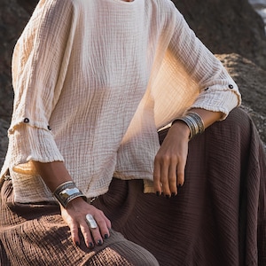 May include: A cream-coloured, long-sleeved top with buttoned cuffs and a brown skirt. The top has a textured, crinkled appearance. Silver bracelets and a ring are visible. The outfit is casual and bohemian in style.