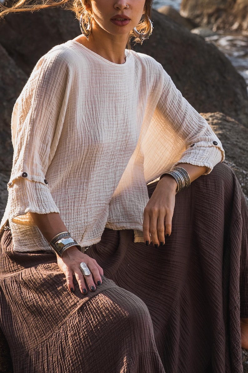 May include: A cream-coloured, long-sleeved top with buttoned cuffs and a brown skirt. The top has a textured, crinkled appearance. Silver bracelets and a ring are visible. The outfit is casual and bohemian in style.