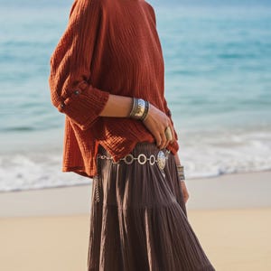 May include: A terracotta-coloured top with long sleeves and a tiered brown skirt. The outfit is accessorized with silver bracelets, rings, and a belt. The background features a beach and ocean.