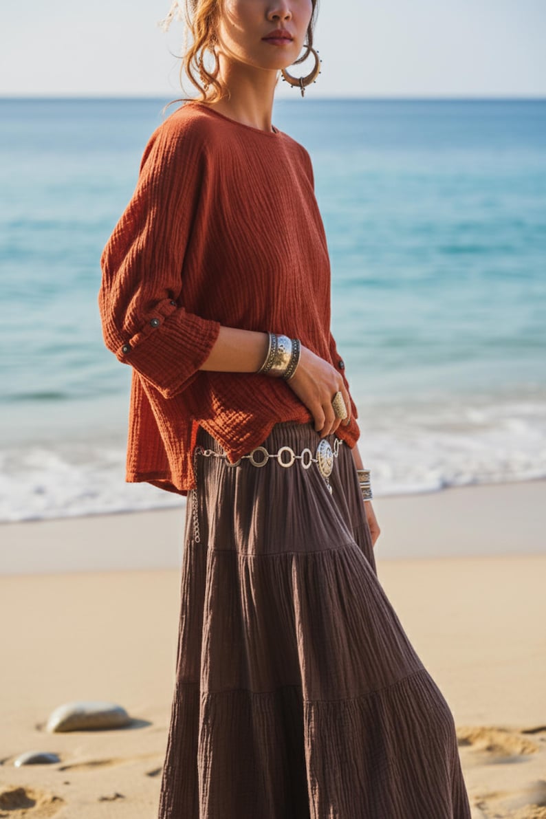 May include: A terracotta-coloured top with long sleeves and a tiered brown skirt. The outfit is accessorized with silver bracelets, rings, and a belt. The background features a beach and ocean.