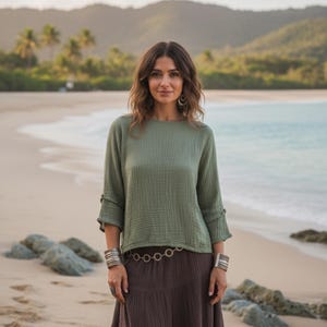 May include: An olive green top with three-quarter sleeves and a brown skirt. The top has a textured appearance and is paired with a long, flowing skirt. Silver bracelets and a belt with circular links complete the outfit. The background shows a beach scene.