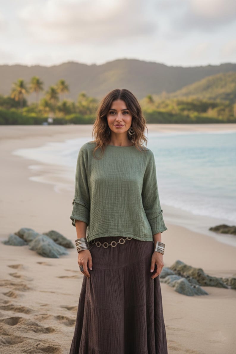 May include: An olive green top with three-quarter sleeves and a brown skirt. The top has a textured appearance and is paired with a long, flowing skirt. Silver bracelets and a belt with circular links complete the outfit. The background shows a beach scene.