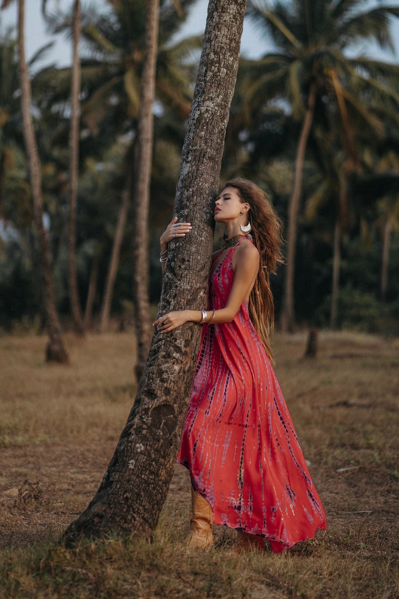 May include: A woman with long, braided hair wearing a red and purple tie-dye maxi dress and brown boots stands next to a palm tree in a tropical setting.