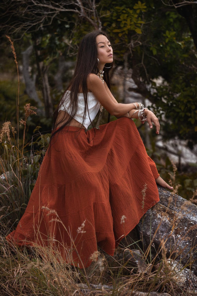 May include: A woman wearing a white crop top and a rust-colored tiered maxi skirt sits on a rock in a natural setting. She is adorned with silver bracelets and necklaces. The skirt has a flowing, textured appearance.