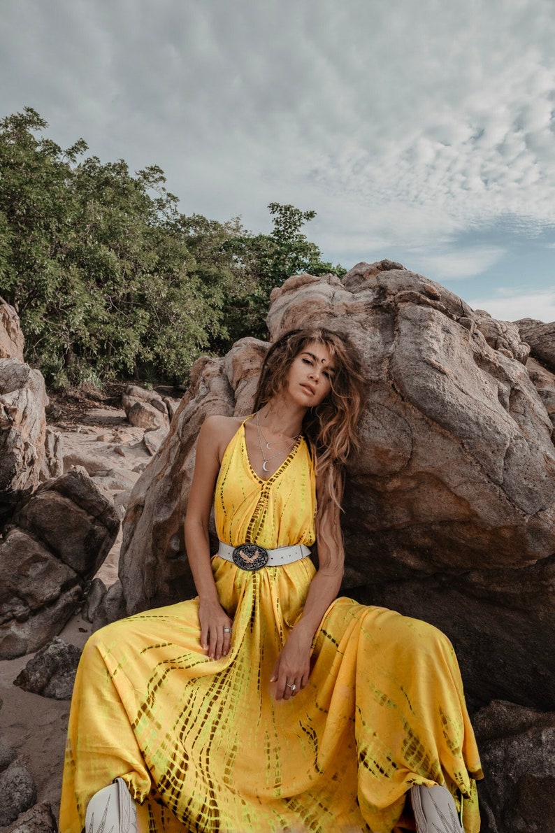 May include: A woman wearing a yellow maxi dress with a white belt and a silver necklace sits against a large rock. The dress has a tie-dye pattern and the woman has long brown hair.