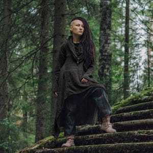May include: A person with short, shaved hair and long dreadlocks is wearing a long, dark gray tunic with a cowl neckline and a pair of plaid pants. They are standing on a set of mossy stone steps in a forest setting.