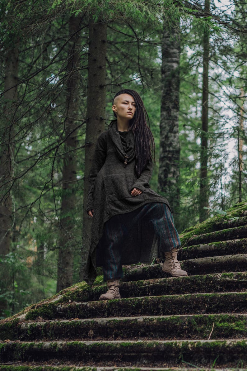 May include: A person with short, shaved hair and long dreadlocks is wearing a long, dark gray tunic with a cowl neckline and a pair of plaid pants. They are standing on a set of mossy stone steps in a forest setting.