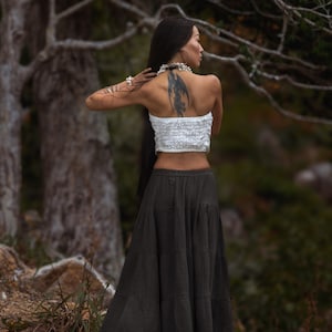 May include: A woman wearing a white tube top and a long, dark green skirt. The skirt is tiered and flowing. The woman has long, dark hair and a tattoo on her back. She is standing outdoors near trees.
