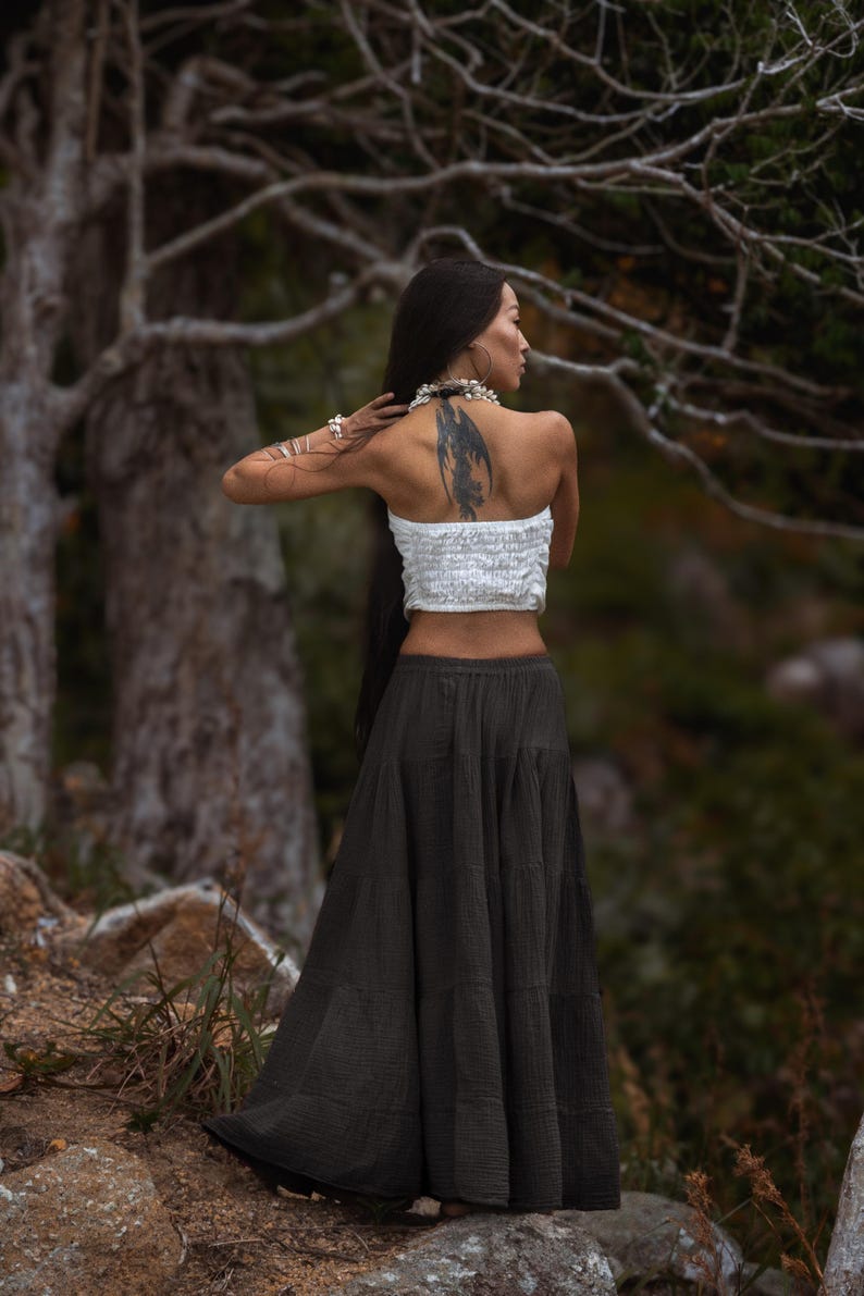 May include: A woman wearing a white tube top and a long, dark green skirt. The skirt is tiered and flowing. The woman has long, dark hair and a tattoo on her back. She is standing outdoors near trees.