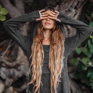 Puede incluir: Una mujer con largas rastas rubias lleva un su&eacute;ter gris con capucha y mangas largas. Se est&aacute; tapando los ojos con las manos, que est&aacute;n adornadas con anillos de plata.