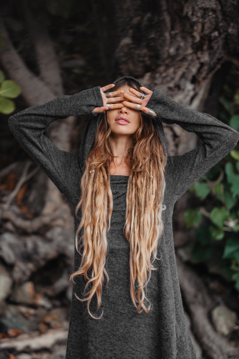 Puede incluir: Una mujer con largas rastas rubias lleva un su&eacute;ter gris con capucha y mangas largas. Se est&aacute; tapando los ojos con las manos, que est&aacute;n adornadas con anillos de plata.