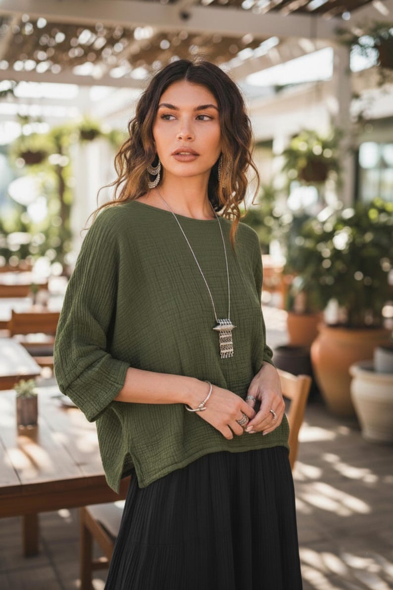 May include: An olive green top with a round neck and long sleeves. The top is worn with a black skirt and silver jewellery, including a long necklace and earrings. The outfit is worn outdoors.