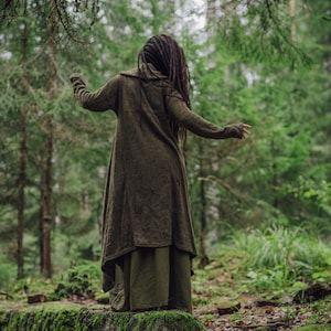 May include: A person wearing a long, dark green cardigan with a hood, standing in a forest. The person has long, dark hair and is wearing a long, dark green skirt.